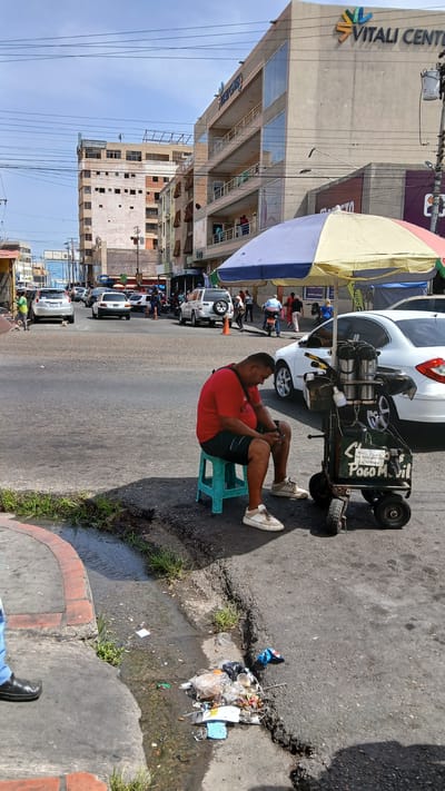 A man sits on a turquoise plastic stool next to a three-wheeled food cart under a striped umbrella on a city street. He wears a red t-shirt and black shorts and is using a mobile phone. The food cart has coffee pots and "Poco Mobil" printed on its side. A white car is parked behind the cart. A pile of trash is visible near the curb. Cars and buildings are visible in the background. The building on the left has "Vitali Center" printed on its exterior. The location is Puerto La Cruz, Venezuela.