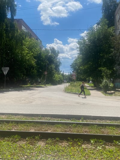 A person is skateboarding down a paved road in Kolomna, Russia. The scene is bathed in sunlight, suggesting it is daytime. The sky is blue with fluffy white clouds, indicating pleasant weather. Lush green trees line the sides of the road, with some buildings visible in the background.
The skateboarder is a young person wearing dark pants and a light-colored shirt. They are in motion, their body angled as if balancing on the board, with one leg extended behind them.
Several traffic signs are visible along the road. One sign shows a red circle with a diagonal line through it, indicating a prohibition, and a vehicle symbol inside. Another sign shows the number "40" within a red circle, likely indicating a speed limit. A triangular sign with a white background and a red border is also present.
The road appears to be a typical urban street, possibly a residential area, with a grassy verge and some overgrown vegetation. The overall impression is a casual, outdoor activity on a clear day.