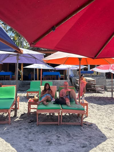 In Karangasem, Indonesia, on a sandy beach, a red umbrella shades a man and woman lounging on green beach chairs. The woman, in the foreground to the left, is wearing a blue and white patterned skirt and a brown top, engrossed in her smartphone. Beside her is a small table with a coconut drink and a bag. The man, to her right, is wearing blue and white striped shorts and is reading a book. A bottle of water and another bag are near him. Behind them, a woman in an orange top and a pink sarong walks past. In the background, tables with blue tablecloths are set up under purple and orange umbrellas, suggesting a beachfront restaurant. Palm trees are visible above the structures, and the sun casts shadows across the sand.