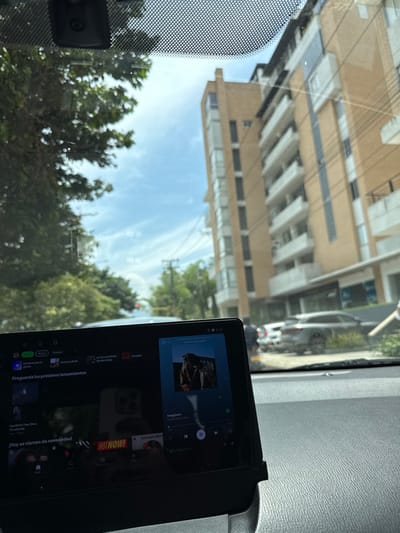 The image presents an urban street scene viewed from inside a vehicle in Pitalito, Colombia, during daytime. The immediate foreground shows a grey vehicle dashboard, on which a black digital screen is mounted. This screen displays a music application interface, prominently featuring the song "Imagínate" by Kendo Kaponi, Cosculluela, and Ñengo Flow, alongside other Spanish language menu options. Reflections are visible on the screen surface. Above the dashboard, the vehicle's windshield is visible, characterized by a dotted pattern at the top and a black dashcam or sensor module centrally mounted.

Through the windshield, a multi-story building with a light brown facade, white balconies, and large ground-floor windows dominates the right side of the view. To the left, lush green trees line the street, partially obscuring a red traffic light further down the road. Utility poles and overhead power lines are visible against a bright blue sky with scattered white clouds. Several vehicles, including a grey SUV, are parked or in motion on the street in front of the building. No individuals are explicitly visible or identifiable.