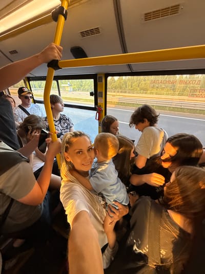 The image depicts the interior of a crowded bus, likely in Borisovo, Russia, given the Cyrillic text visible on a sign that reads "ЗАПАСНЫЙ ВЫХОД" (Emergency Exit). Several individuals are visible, with a woman in the foreground holding a child. The bus has yellow handrails and windows showing a landscape with trees and a road. The lighting suggests it is either sunrise or sunset, casting a warm glow on some of the passengers. People are seated and standing, with some holding onto the handrails for support. The overall atmosphere appears to be one of travel in a public transportation vehicle.