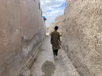 A man in a long, brown, loose-fitting garment walks down a narrow alleyway between two high, earthen walls in Saranan, Pakistan. The walls are both made of rough, unplastered mud bricks, and there are some white scribbles on the wall to the left. The ground is dirt and dusty.  The man's back is to the camera and he appears to be in his mid-twenties. He is wearing a dark-colored cap. The man's right hand is in his pocket and his left hand is swinging at his side. The alleyway is narrow, not more than five feet wide, and the walls are high, creating a sense of enclosure.  The sky is overcast with a few wispy clouds visible.  The scene is quiet and peaceful.  The coordinates 30.532, 66.909, place the image in a relatively rural part of Saranan.