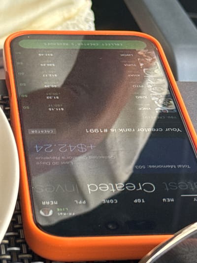 The image shows a close-up of a smartphone with an orange case, resting on a dark, woven surface. The screen displays a dashboard or profile related to a creator, showing statistics like "Total Memories: 503," "Your creator rank is #1991," and "Collected Creator's Revenue: +$42.24." There are also lists of names and associated monetary values, such as "$11.15," "$1.32," and "$6.39," with some indicating fractional amounts. Text labels like "NEW," "TOP," "CORE," "PPL," and "LIVE" are also visible, suggesting different categories or statuses within the platform. The overall impression is one of a digital interface presenting performance metrics for a content creator. The lighting and the angle suggest the photo was taken outdoors during the daytime, possibly in a cafe or restaurant setting, given the outdoor furniture texture. The reflection on the screen hints at the surrounding environment, but no people are clearly depicted. The context of "la Vila Joiosa, Spain" suggests a coastal or Mediterranean location, though this is not directly evident from the image itself.