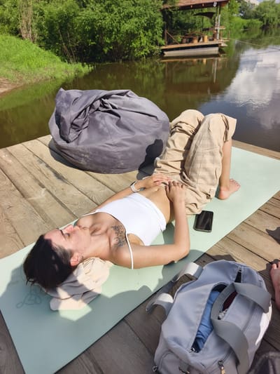 A woman is lying on a yoga mat on a wooden deck next to a body of water. She is on her back with her eyes closed, wearing a white tank top and light-colored pants. Her arms are resting on her stomach. Next to her is a smartphone and a gray duffel bag. A large gray beanbag chair is to her left. In the background, there is lush green foliage and a wooden structure on the water. The scene appears to be in a natural setting near Онохино, Russia.
