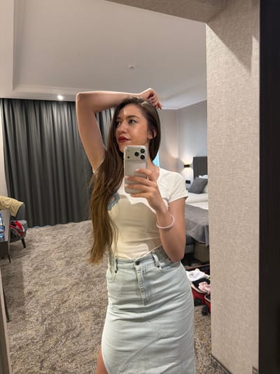 A young woman with long brown hair and bold red lipstick is taking a mirror selfie in what appears to be a hotel room. She is wearing a white t-shirt and a light blue denim skirt. Her right arm is raised, and she is holding a smartphone in her hand, which is angled to capture her reflection. A pink bracelet is visible on her wrist. The background of the mirror shows a bed with grey bedding, and a glimpse of clothing items, including a red backpack, is visible on the floor. The room has textured wallpaper and a carpeted floor. The lighting suggests it is daytime. The woman’s expression is somewhat playful or curious. The scene is set in Hisarya, Bulgaria.