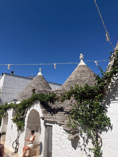 In Alberobello, Italy, under a clear blue sky, a woman in a white dress sits in an arched alcove of a traditional white trullo building. The trullo, characterized by its conical stone roof and white stone construction, is adorned with green ivy. String lights with exposed bulbs hang across the scene. The woman is holding a glass and looking down. The image captures a bright, sunny day with strong shadows cast by the architecture and foliage.