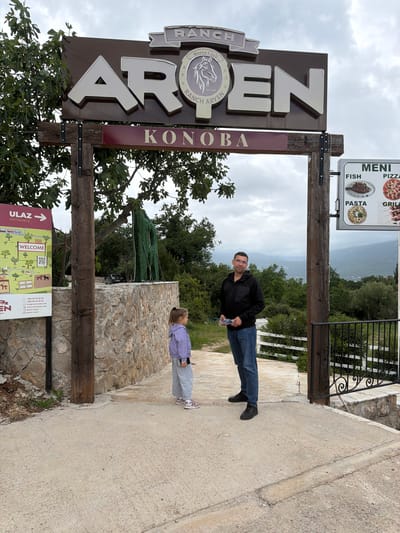 The image depicts a man and a child standing near the entrance sign of "Ranch Aryen KONOB", presumably located in Provodina, Montenegro. The sign is made of wood and metal and features the ranch's name, logo, and restaurant offerings: fish, pizza, pasta, and grill. A menu board is attached to the upper right of the entrance. The surrounding environment includes trees, a stone wall, and a pathway. A smaller sign below the main sign has a map and the words, "ULAZ ENTRANCE," "WELCOME," and various contact details. The image is taken from a slightly rotated perspective.