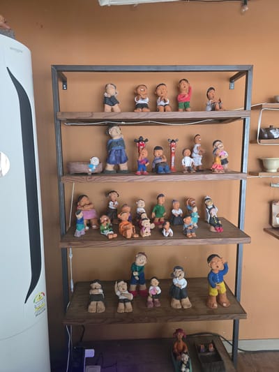 This media file depicts a display of numerous ceramic figurines, primarily of children, arranged on tiered wooden shelves against a textured orange wall. The figurines are diverse in pose and attire, with many depicted in lying or sleeping positions, some with their eyes closed and others appearing relaxed. Some figures are dressed in traditional Korean clothing, while others wear more modern outfits.

The figurines are predominantly human-like, with stylized features and earthy tones, suggesting they are handmade. Several are gathered in groups, while others stand alone. Notable details include some figures holding small objects like hearts or what appear to be books. One figurine at the bottom left is larger and dressed in a blue garment, sitting on a brown, block-like object.

The setting appears to be indoors, possibly a shop or a home display area. The lighting is uniform, suggesting it's not daylight-specific. There are no obvious weather cues or strong emotional expressions on the figurines, which are mostly neutral or serene.

Visible text includes the brand name "WHISEN" on a white appliance in the foreground, which is partially cropped. There are also some labels and symbols on the appliance, including energy efficiency ratings. The location context is Gumi-si, South Korea. The overall impression is one of a collection of decorative folk art or cultural artifacts.