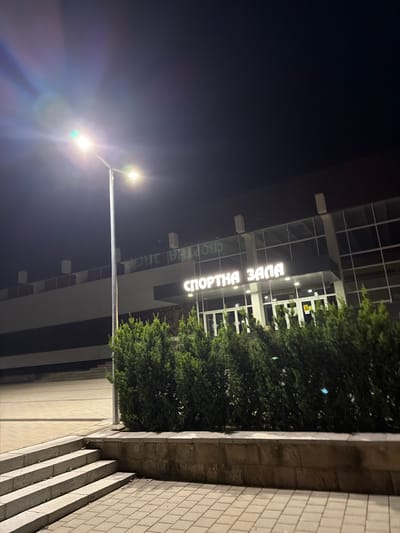 The image shows the exterior of a sports hall at night in Pravets, Bulgaria. The hall has large glass windows and illuminated signage that reads "СПОРТНА ЗАЛА" (Sports Hall). A tall street lamp with two lights illuminates the foreground, casting lens flares. In front of the building is a row of manicured evergreen bushes, and to the left, there are stone steps leading up to a paved area. The overall environment suggests an outdoor public space at night.