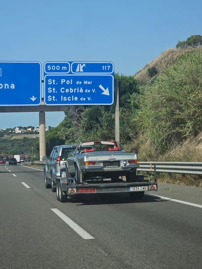 The image shows a highway scene with traffic. In the foreground, a silver SUV is towing a trailer carrying a vintage silver convertible car. The trailer has a license plate that reads "1835 CMB". Behind the SUV, other vehicles are visible on the road. Overhead signs indicate directions to "Sant Pol de Mar" and "Sant Cebrià de V." and "Sant Iscle de V." with distances of "500 m" and "117". The environment is characterized by a clear blue sky and a grassy, wooded hillside to the right of the highway. The general location appears to be the coastal region of Spain, specifically near Sant Pol de Mar.