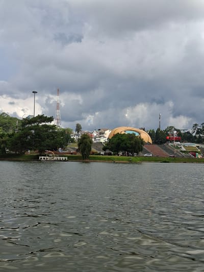 A wide shot captures Xuan Huong Lake in Da Lat, Vietnam, under a dramatic, cloudy sky. The lake occupies the foreground, its surface rippling with greyish-green water. On the far bank, a large, lotus-shaped building with a golden roof dominates the scene, its tiered entrance flanked by numerous red flags and stairs. To the left of the building, a cluster of green trees stands beside a tall, red and white transmission tower and a much taller, grey lamppost. In the mid-ground, various buildings and trees are visible, with a small pier in the lower left corner featuring a large, circular metal sculpture. Several people are scattered around the area, some walking on the steps leading up to the building, others near the lake's edge. A white car and some motorcycles are also present on the paved areas. The overall atmosphere is overcast and somewhat somber due to the heavy clouds.