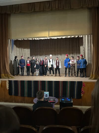 A wide-angle shot shows a theatrical performance in what might be the town hall of Voiniagovo, Bulgaria. A group of roughly a dozen people is standing on stage wearing traditional Bulgarian costumes. The stage has a simple setup with beige curtains on either side and at the top.
To the right of the stage, a black carpet with colorful stripes is seen. A person is sitting in the audience, operating sound equipment. Rows of wooden seats can be seen in the foreground. The location is indicated by a sticker with the town's name on the wall.