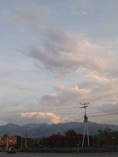 The image presents a view of Parroquia Ocumare, Venezuela, with mountains forming the horizon under a sky filled with numerous clouds. Electrical power lines and support structures are visible in the midground. Along the lower portion, a road stretches horizontally, with a black SUV driving on it. A pedestrian is also present on the roadside. Red and green vegetation is apparent, adding color to the scene. A building or structure is seen on the left side of the image.