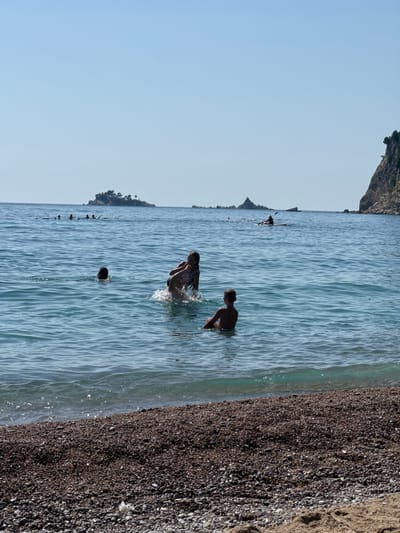 The image shows a beach scene in Đurovići, Montenegro. Several people are in the clear blue water, with some swimming and playing. In the foreground, a boy is sitting in the water, facing away from the camera. To his left, another person is partially submerged, with their head and shoulders visible. Further out in the water, a group of people are seen in the distance, with some appearing to be swimming or floating. A lone kayaker is visible on the water in the middle ground, paddling towards the right of the frame. In the background, small islands with trees are dotted across the horizon, and a rocky cliff face dominates the right side of the image. The beach itself is covered in pebbles, with a hint of sand in the lower right corner. The sky is clear and blue, indicating a sunny day.
