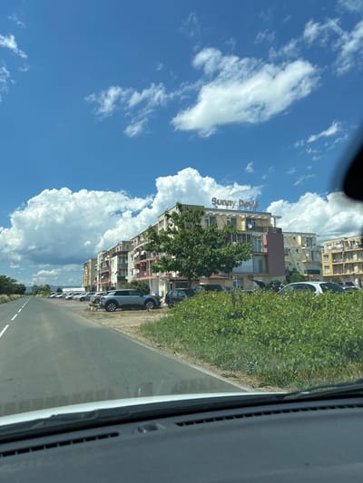 The image shows a street scene in Tankovo, Bulgaria, on a sunny day with scattered white clouds. On the right side of the image, there is a row of multi-story apartment buildings with balconies and satellite dishes. One building has a sign that reads "Sunny Day 5". A large green tree is positioned in front of the buildings. In front of the buildings, there is a parking lot with several cars. The left side of the image shows a paved road with white lane markings. Lush green vegetation grows along the side of the road and in front of the buildings. The photo appears to be taken from inside a vehicle, with the dashboard visible in the foreground.