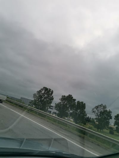 The image captures a view from a vehicle traveling on a highway, likely on the outskirts of Kazichene, Bulgaria. The sky is overcast with thick, gray clouds, suggesting impending or recent rain. The highway itself is paved with asphalt, marked by a white line on the right side. A metal guardrail separates the road from the adjacent grassy area and a row of trees. Beyond the trees, several modern, large, box-like buildings with gray facades and white window frames are visible, indicative of industrial or commercial structures. Some trees are in the foreground and middle ground, appearing green and full. A white truck is partially visible on the far left of the frame, suggesting ongoing traffic. The overall scene is one of a utilitarian, functional landscape with a muted color palette dominated by grays and greens, under a somber sky.