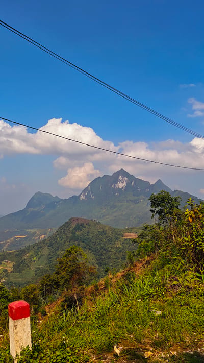 The image depicts a landscape in Mậu Duệ Commune, Vietnam, featuring mountains, a partially cloudy blue sky, vegetation, and infrastructure. The focal point is a range of mountains in the background, with the largest peak exhibiting exposed rock faces on its slopes. Green, forested vegetation covers the lower portions of the mountains. In the foreground, a roadside embankment is covered with lush green grasses and various plants. A red and white concrete post stands on the embankment's edge. Two power lines run diagonally across the upper portion of the sky. The sky is mostly blue with scattered white cumulus clouds. Patches of land with terraced agriculture are visible in the mid-ground, indicating human modification of the landscape. The scene captures a combination of natural mountainous terrain with elements of human intervention, such as power lines and agricultural land use.