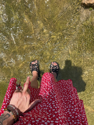 A top-down shot depicts a person standing in shallow water. The water appears clear with an olive-green tint, revealing the riverbed beneath. The person is wearing a red dress with a small white floral pattern and black open-toed sandals. One hand is visible, holding the edge of the dress and pointing down towards the water with the index finger. The wrist has bracelets and a tattoo is visible on the arm. The scene is located in Picassent, Spain.