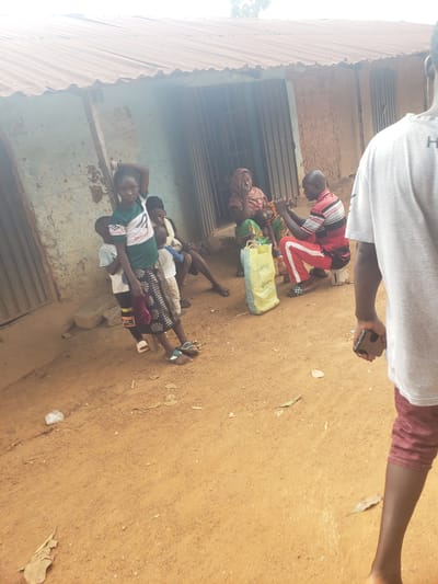 In Kachia, Nigeria, a group of individuals are gathered outside a structure with a corrugated metal roof and mud walls. In the foreground, a person wearing a grey t-shirt and maroon shorts is seen from the back, holding a mobile phone. To their left, a young girl in a green t-shirt and patterned skirt stands, holding a cloth. Behind her, two young boys and a woman are partially visible. Further back, a man in a striped shirt and red pants is seated, and another woman in a headscarf is also present. A yellow plastic bag and a water bottle are on the ground near the seated man. The environment appears to be a dusty, outdoor area.