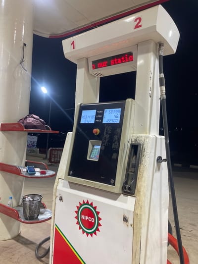 The image shows a gas pump at a NIPCO fuel station in Guzape, Nigeria. The gas pump is white with red and green accents. The digital display shows a price of 403, a liter count of 4.80, and a total cost of 1229.00. A red sign above the display reads "our statio" in red LED lights. The pump has a black hose attached and a number "1" on the front. To the left of the pump are shelves with various items, including a phone, a small trash can, and a water bottle. The ground is paved. The scene appears to be at night, with artificial lighting visible.
