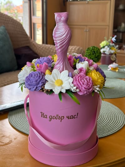A floral arrangement in a pink cylindrical box is displayed on a wooden table. The box has a pink ribbon handle and text written in gold script on its side that reads "На добър час!" which translates to "Good luck!". The arrangement itself features an assortment of artificial flowers in various colors, including purple, pink, yellow, and white, with a prominent pink, twisted dress-shaped ornament at the center. The background suggests an indoor setting with a window, part of a wicker chair, and wooden cabinetry. The lighting indicates it is daytime. The text on the box is in Bulgarian, a language spoken in Lyubimets, Bulgaria, but the specific location within the city is not discernible from the image alone.