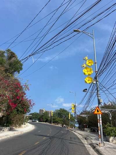 The scene depicts a street in Nam Nha Trang, Vietnam, characterized by a dense overhead tangle of electrical wires against a clear blue sky. A streetlamp with yellow flower decorations hangs among the wires. A yellow and black road sign is visible. The road curves slightly uphill, marked with dotted yellow lines. Red and green foliage lines the roadside. A building is visible in the distance beyond trees. The image is taken from a slightly low angle, emphasizing the verticality of the wires and streetlamp.