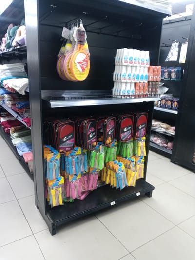 This image depicts an aisle within a retail store located in Kawaji, Nigeria. The scene is dominated by a tall, black metal shelving unit in the foreground, displaying various consumer goods. To the immediate left, a partial view of another shelving unit shows stacks of folded textiles, including neck pillows in shades of teal, beige, and purple on the top shelf, and folded towels or clothing in white, blue, green, and maroon on lower shelves.
The main black shelving unit features three distinct display levels. At the top, a series of empty hanging hooks are present against a black panel. Below this, multiple mesh bags each containing a badminton set (two rackets and two shuttlecocks) are suspended from hooks; the rackets are brightly colored in yellow, pink, and orange. The middle section of the unit consists of a flat shelf, where multiple stacks of white plastic bottles with blue labels are arranged on the left, next to several stacks of brown-orange bottles of similar size and shape on the right. Behind these bottles, packages appearing to contain men's undergarments are visible. The lower section of the shelving unit features numerous packaged jump ropes hanging from hooks, displayed in assorted colors including blue, green, yellow, and pink. Each package is labeled "JUMP ROPE" and indicates a counter feature on the handle. Behind the jump ropes, multiple red and black cases, branded "FUKANG RACKET" and likely containing table tennis paddles, are arranged vertically. The bottom shelf of this unit displays additional jump ropes and has white price tags affixed to its edge, some showing "1.00".
To the right, a segment of another black shelving unit is visible, holding items in plastic packaging, including what appears to be folded clothing or undergarments. The floor throughout the visible area is covered in light-colored square tiles with dark grout lines. No individuals are present in the scene.