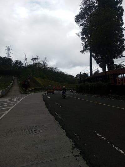 The image presents an overcast daytime scene on a paved road in Deothang, Bhutan. The dark asphalt road, featuring white dashed lane markings and a continuous yellow line, curves slightly to the right and recedes into the distance. A motorcyclist, wearing a light-colored helmet and dark clothing, operates a dark motorcycle in the midground, traveling away from the viewer. Ahead of the motorcycle, a maroon van proceeds in the same direction, followed by a lighter-colored vehicle further in the distance.
To the immediate left of the road, a grey concrete pedestrian path with white diagonal striped markings transitions into a set of ascending concrete steps alongside a steeply rising, vegetated hillside. A circular red speed limit sign, displaying "11" in white, is mounted near the base of the hill. Further up the hillside, which is covered with dense green foliage and several bare trees, a metal lattice-work electricity transmission tower with visible power lines stands prominently against the cloudy sky.
On the right side of the road, a dense line of tall evergreen trees obscures parts of the background. Behind these trees and a lower green hedge, a building with a reddish-brown tiled roof and dark walls is partially visible; large dark letters are discernible on its facade. Overhead power lines are also present on the right side of the scene. The sky is uniformly overcast with light grey clouds.