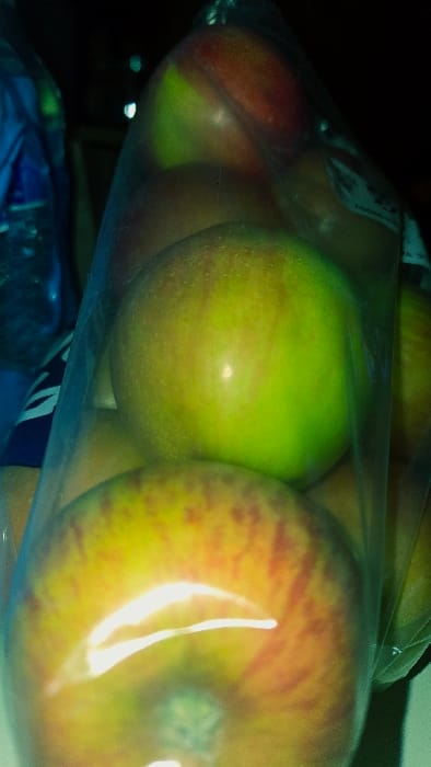 The image displays a close-up, vertical view of multiple bi-colored apples contained within a clear, flexible plastic bag. Approximately seven apples are visible, exhibiting skin tones of green and reddish-orange, with some yellow mottling. Prominent bright reflections are present on the surfaces of the apples and the plastic bag, indicating a direct light source. To the left of the bag, a blurred blue plastic object, likely a bottle, is partially visible, showing faint white markings. The background is dark and out of focus. No individuals, specific actions, or interactions are depicted. The scene is located in Lompoc, United States.