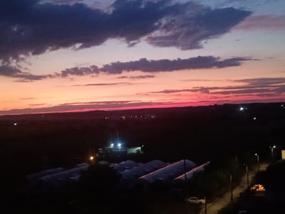 The image depicts a twilight sky over a dark, rural landscape. The sky displays a gradient of colors, transitioning from deep purple and blue clouds at the top to vibrant pink and orange hues near the horizon. Below the sky, the ground is predominantly dark, with some scattered lights visible, suggesting a sparsely populated area or distant settlements. In the foreground, several long, low-lying structures, possibly greenhouses or agricultural buildings, are visible. Their reflective surfaces catch some of the ambient light. A paved road with streetlights runs through the scene, with a white car parked alongside it. The overall impression is one of a quiet evening scene in a rural setting, likely on the outskirts of Aidemir, Bulgaria.