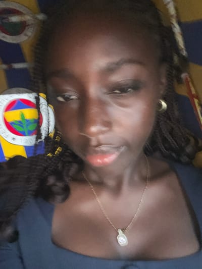 A dark-skinned person with braided hair is lying on a bed with a Fenerbahçe Spor Kulübü can or mug. The person wears a gold necklace and earring. The location is potentially in Galadimawa, Nigeria.