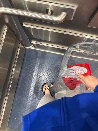 A high-angle shot depicts a person ascending stairs, possibly an escalator, in a public building. The person is wearing a blue hoodie, grey sweatpants, and black flip-flops. They are carrying a grey mesh shopping bag containing a red and white shopping bag. The stairs/escalator features a metallic handrail and a blue, textured surface with circular patterns. The scene is illuminated by bright, diffused light. The location appears to be within a shopping centre or transportation hub. The city of Hull, United Kingdom, cannot be verified through the image.