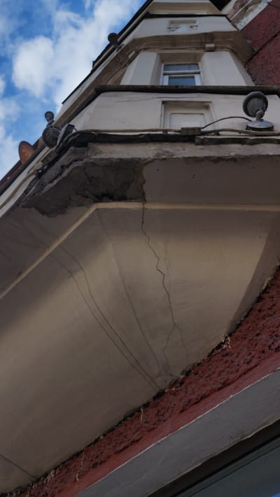 The image shows an upward-angled view of a building's exterior. The architecture is characterized by a mix of off-white and grey painted surfaces, with a section displaying exposed brick. Cracks and deterioration are visible in the plaster, particularly along the lines of the building's structure. Electrical conduit and lights are mounted on the exterior walls. The sky with scattered clouds is visible in the lower left of the frame, indicating an outdoor setting. Based on architectural style and given the prompt, this likely depicts a building in Smolensk, Russia.