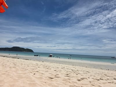 This media file depicts a serene beach scene in Bang Sare, Thailand. The foreground is dominated by a sandy beach with gentle ripples and footprints, suggesting a low tide or recent activity. In the middle ground, the clear, turquoise ocean meets the shore, with several small boats and a few people scattered along the waterline. One boat appears to be a speed boat with people on board, while another is a smaller inflatable vessel. A couple of individuals are wading in the shallow water, and a lone person is walking along the wet sand. In the distance, a lush green island rises from the sea, its slopes covered in dense vegetation. The sky above is a vibrant blue, streaked with wispy white clouds, indicating a bright, possibly mid-morning or early afternoon, day with pleasant weather. The overall atmosphere is one of tranquility and relaxation, typical of a tropical beach getaway. A small orange object, possibly a flag or banner, is visible in the upper left corner of the frame, partially obscuring the sky. No visible text is present in the image.
