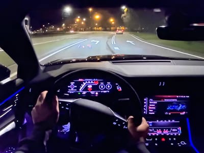 The image shows a first-person view from the driver's seat of a car at night. The car is driving on a road with streetlights and trees visible in the background. The dashboard displays various information on a digital screen, including speed (48 km/h), navigation arrows, and other vehicle data. The steering wheel is visible, with a person's hands on it. The car's interior is illuminated by ambient lighting. There are no discernible landmarks that would indicate the location of Bezden, Bulgaria.