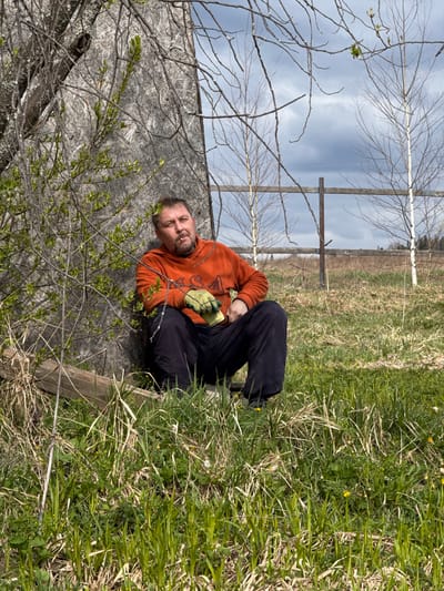 A middle-aged man is lying on a sheet of plywood propped against a fence in a grassy field. He is wearing an orange sweatshirt, dark pants, and gloves. He appears to be smoking. The scene is outdoors, possibly near the city of Усть-Тунтор, Russia, based on geographic context. The background includes a fence, trees, and a field.