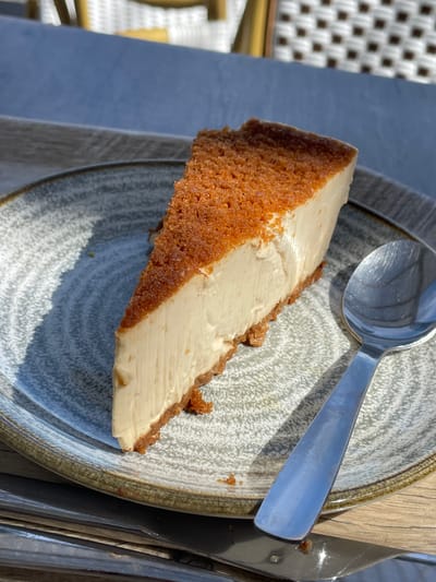 A slice of cheesecake sits on a decorative plate. The cheesecake has a thick, brown crust and a creamy, pale filling. A silver spoon rests on the plate, angled towards the top right of the image. To the right of the plate, a metal fork is visible. The plate is placed on a wooden surface, and in the background, a woven white chair is slightly out of focus. The lighting suggests it is daytime, with a bright light casting shadows. The scene appears to be an outdoor setting, possibly a cafe or restaurant patio, given the woven chair and outdoor elements. No text is visible in the image.