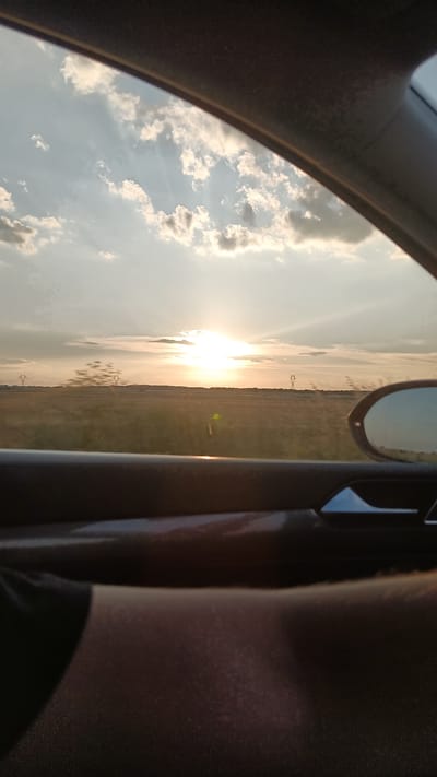This is a photograph taken from inside a vehicle, looking out the window towards a sunset over a rural landscape. The sun is visible in the sky, partially obscured by clouds, casting rays of light across the scene. The landscape appears to be a field or farmland with some utility poles in the distance. The interior of the car is visible in the foreground, with a door handle and part of a person's leg. The photograph was taken in the vicinity of Radievo, Bulgaria.