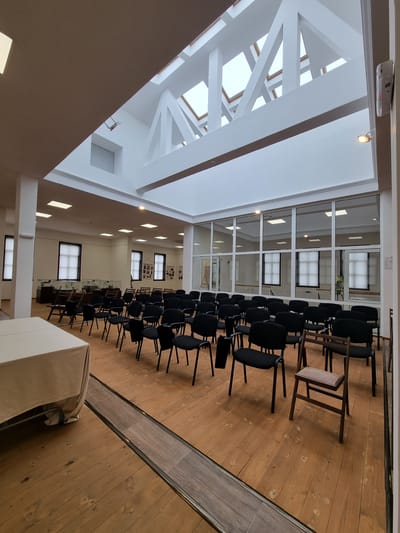 The image depicts an interior space in the city of Elena, Bulgaria, likely a public hall or auditorium. Rows of black chairs are arranged on a wooden floor, suggesting a setup for an audience or presentation. Some wooden chairs are interspersed among the black ones, adding to the seating. Windows are visible along one side of the room, providing natural light. The ceiling features a modern, white-painted framework, possibly supporting a skylight. The room appears empty, devoid of people, indicating it's either pre- or post-event. A covered table, likely for speakers or displays, stands in the foreground.