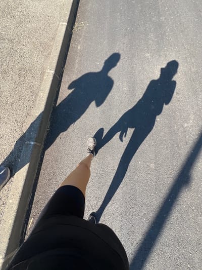 The image depicts an outdoor street scene in Puyloubier, France, under direct sunlight, indicated by long, distinct shadows. The ground surface consists of dark asphalt, bordered on the left by a concrete curb that separates it from a lighter, coarse-textured paved area. In the immediate foreground, the lower torso, upper right leg, and right foot of a person are visible. This individual is wearing dark (likely black) athletic shorts and a light-colored athletic shoe with dark accents on the right foot. Three elongated human shadows are cast across the asphalt. One shadow originates from the individual holding the camera, extending from the bottom right of the frame towards the upper left. To the left of this shadow, two additional humanoid shadows are visible: one appearing to be a male figure, with arms positioned across the chest or at the sides, and another appearing to be a female figure. The presence and length of the shadows suggest multiple individuals are walking or standing on the thoroughfare, with the sun at a low angle.