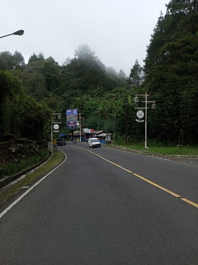 The image captures an overcast, possibly foggy, day on a winding asphalt road, situated in the city of Phoxay, Laos. Dense, dark green vegetation, including coniferous trees, covers the steep hillsides on both sides of the road, with mist obscuring higher elevations in the background. The road surface is dark grey, with a solid white line marking the left edge and a broken yellow line separating traffic lanes. A concrete drainage channel runs alongside the left curb.

In the mid-ground, a white panel van travels away from the viewer in the right lane. Further down the road on the left, near a collection of small roadside stalls, a dark-colored pickup truck is visible, followed by a motorcycle. The stalls exhibit simple construction with light-colored walls and corrugated roofs. A prominent gateway structure spans the left side of the road, featuring a large blue banner advertising "Garden" and "2J Puncak," flanked by two decorative white clock faces. On the right side of the road, a white decorative lamppost stands, adorned with three ornate lanterns and a circular sign displaying Arabic script for "Allah." A standard street light is partially visible in the upper left foreground. Utility poles carrying wires are present along both sides of the road. No individuals are clearly discernible.