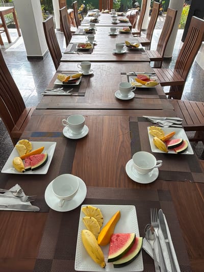A long wooden table is set for breakfast in Rumassala, Sri Lanka, with individual place settings featuring fruit platters and teacups. The table is set with white square plates displaying an assortment of sliced fruits including watermelon, pineapple, mango, and banana. Each setting also includes a white teacup and saucer. Cutlery, consisting of a knife, fork, and spoon, are placed to the left of each plate, resting on a napkin. Wooden chairs with slatted backs are positioned along the table, with the chairs on the right appearing more in focus. The floor is tiled with a reflective surface. The background shows glimpses of outdoor greenery and a yellow motorcycle parked in the distance, suggesting an open-air or semi-outdoor dining area. The overall scene is tidy and prepared for a meal.