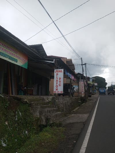This image depicts a street scene in what is identified as Ariyalai East, Sri Lanka, on an overcast day.
On the left, a multi-tiered concrete retaining wall supports a raised commercial area. The wall is partially covered in green vegetation, primarily grass and small plants, with some litter visible. A set of four concrete steps ascends the wall from the lower grassy verge. Above the retaining wall, two adjacent single-story commercial structures with corrugated metal roofs extend along the street. The leftmost structure, partially visible, displays a yellow and orange banner with red text, partially legible as "DAN INTERIOR" and "PARTAI BESAR DAN ECERAN" (meaning "and interior," "wholesale and retail" in Indonesian), along with a partial phone number "5267181698". Inside this structure, a single individual is seated on a light blue stool behind a desk or counter, with various long, narrow materials, possibly building supplies, stacked vertically nearby.
To the right of the first structure, a white rectangular banner with prominent red text reading "JUAL PLAFON PVC" (meaning "Sell PVC Ceiling" in Indonesian) and a phone number "0852-6718-1698" hangs vertically from the building's overhang, facing the road. Under this overhang, additional items are stored or displayed, and several colorful garments are strung on clotheslines. A dark-colored pickup truck is parked on the raised platform, facing right, partially obscured by the "JUAL PLAFON PVC" banner.
The right side of the image features a two-lane paved road, stretching into the distance and slightly uphill. A solid white line marks the road's edge on the right, and a broken white line is visible further down, delineating the center of the road. In the mid-distance on the road, two blue utility vehicles, possibly small trucks or SUVs, are either stationary or moving away from the viewer. A third dark-colored vehicle follows the second blue vehicle. Multiple utility poles with a dense network of overhead wires crisscross against the sky.
The sky is heavily overcast with dense gray clouds. Trees are visible in the background, to the right of the road, along with additional distant buildings and structures lining the street.