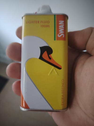 A person is holding a rectangular tin of Swan Lighter Fluid. The tin is predominantly yellow with a white swan graphic on it. Red text on the tin reads "LIGHTER FLUID" and "100ML". To the right of the swan graphic, the word "SWAN" is written vertically in red letters within an orange rectangle. The bottom edge of the tin has red text that reads "EXTRA REFINED". The background is a blurred, light-colored surface, possibly a wall or ceiling. There is no indication of location within Huntingdonshire, United Kingdom.