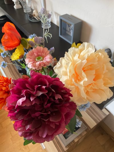 This media file captures a close-up, top-down view of a vibrant artificial flower arrangement. The arrangement features several large, layered flowers in shades of pale yellow, deep magenta, and a soft pink, resembling peonies and possibly a gerbera daisy. Smaller accent flowers in blue and yellow are interspersed throughout the bouquet, along with delicate sprigs of lavender. A bright orange poppy and an orange mum are also visible.
The flowers are arranged in what appear to be clear glass vases, one of which is partially obscured by other elements in the foreground. Behind the flowers, a dark surface, likely a table, is visible. To the left, a small, grey, box-like object with a transparent window is present. Inside this object, a piece of paper with text is visible, including the German word "Reisezeit," meaning "travel time."
The background shows a mix of surfaces. On the left, there is a textured, off-white wall. To the right, a wooden surface with a striped pattern and another wooden surface that appears to be a floor or tabletop are present. The lighting suggests it is daytime, with soft light illuminating the scene. There are no people visible in the image. The overall impression is one of a decorative still life, likely set indoors. The presence of "Reisezeit" on the paper hints at a theme of travel or holidays. The location context of Lütjenwestedt, Germany, provides a geographical setting for this scene.