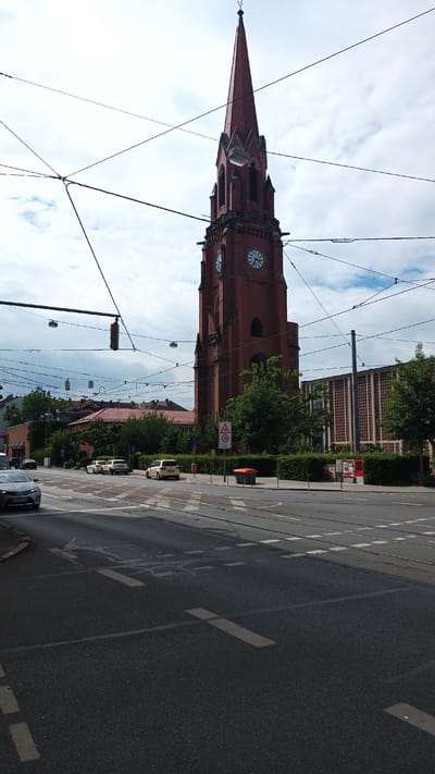 The image shows a street scene in Nuremberg, Germany, featuring a prominent red brick church tower with a tall, pointed steeple. The tower has clock faces on at least two sides. The sky is overcast, suggesting a cloudy day. Overhead, numerous electrical wires, likely for trams or trolleys, crisscross the frame, obscuring parts of the sky and the church.
In the foreground, a wide paved street with tram tracks is visible. Several cars are on the road, and there are crosswalk markings. The street is bordered by a sidewalk with a neatly trimmed hedge. A speed limit sign showing "30" with a red circle around it is present. To the right of the sign, a modern building with a grid-like facade is visible behind the hedge.
A few trees are scattered throughout the scene, with more buildings in the background, some with red tile roofs. The overall impression is of a typical urban environment with a blend of historic architecture (the church) and modern infrastructure. There are no people clearly visible in the image, and the scene appears to be during daylight hours. The mood is neutral, conveying an ordinary day in the city.