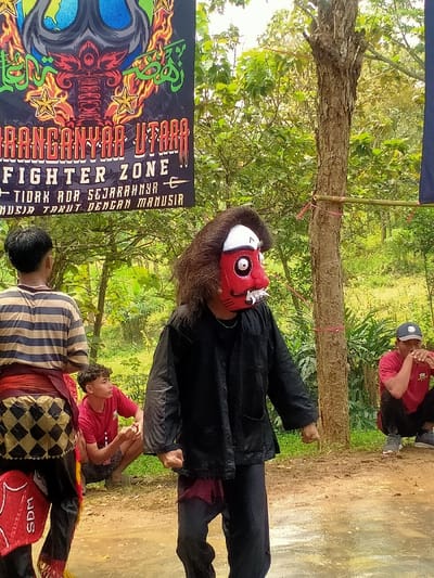 An outdoor scene in Tamansari, Indonesia, depicts a performance or gathering involving four males on a reddish-brown, possibly damp, dirt ground. The central figure, facing slightly right, wears a red mask with white eyes, prominent fangs, and a white forehead design, framed by shaggy dark hair. This individual is dressed in a black long-sleeved jacket over a dark undershirt, dark pants, and features a visible dark red fabric under the jacket hem; fists are clenched. To the left, a figure with their back to the camera wears a striped brown and beige shirt, a black and gold checkered sarong-like garment, dark pants, and a red shin guard with "SDM" in white. In the left-middle background, a male in a red t-shirt and dark shorts crouches, observing the masked figure. To the right, another male in a red t-shirt and dark pants, wearing a blue baseball cap, squats with clasped hands, also observing. Behind the subjects, a dark banner with white and red text reads "ARANGANYAR UTARA", "FIGHTER ZONE", and "TIDAK ADA SEJARAHNYA MANUSIA TAKUT DENGAN MANUSIA" (No history of humans fearing humans), featuring stylized flames, stars, and a trident-like symbol. Dense green foliage and tree trunks form the background, with a prominent tree trunk to the right of the masked figure, to which a bamboo pole is horizontally tied with red string.