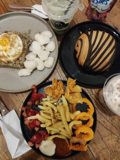 A top-down, medium shot captures a rustic wooden table laden with a meal. To the left, a grey plate holds a portion of fried rice topped with a sunny-side-up egg and accompanied by a pile of white crackers. Next to it, a black plate presents a pastry with dark chocolate stripes, possibly a danish or croissant. In the foreground, another black plate displays a hearty serving of french fries, crispy onion rings, and what appear to be deep-fried wontons and cut hot dogs. Two small dollops of sauce, one red and one white, are also on this plate. To the right, a clear plastic cup contains a green beverage with ice and a straw, and a clear plastic cup further to the right holds a creamy, frothy drink topped with some garnish. A small packet with the word "Bites" and strawberry imagery is visible behind the green drink. A white napkin is partially visible on the left edge of the frame. The overall scene suggests a casual dining experience, possibly at a cafe or restaurant in Pagerwojo, Indonesia, given the presence of local Indonesian crackers. No people are visible in the image.