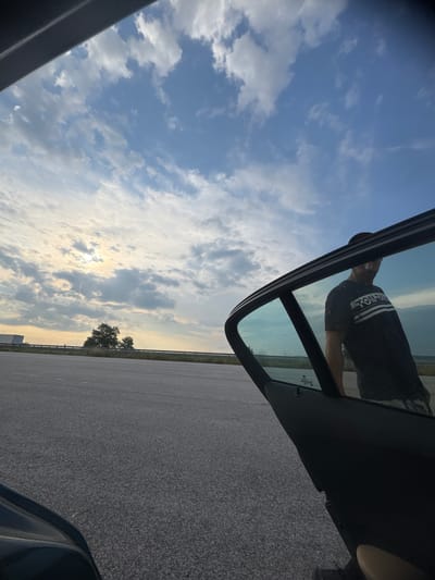 The image depicts an outdoor scene in Rupkite, Bulgaria, at what appears to be a roadside rest area or parking lot during late afternoon or early evening. The upper portion of the frame shows a partially cloudy sky with a bright sun low on the left horizon, casting a strong light through scattered cumulus clouds. The ground consists of dark gray asphalt pavement, extending into the midground. Along the distant horizon, a metal guardrail is visible, with sparse green vegetation and a cluster of trees directly beneath the brightest part of the sky. A large white semi-trailer truck is partially visible on the far left side of the horizon line.
In the foreground and midground on the right, the open front door of a vehicle is prominently displayed, viewed from the car's interior. The car door, including its frame and interior panel, is dark-colored. The door's window is tinted and reflects a male figure and portions of the cloudy sky. The reflected male figure, seen from the upper torso, wears a dark t-shirt with white horizontal stripes and partially legible text that reads "TOM TAN" or similar. His face is somewhat obscured by the reflection and angle, and he appears to be looking towards the left side of the frame. Within the reflection on the car window, a sticker bearing the text "MITSUBISHI MOTORS" is visible on the glass. Faintly visible on the interior window frame is a subtle "BMW" marking. A light tan/beige material, possibly part of the car's interior seating or floor, is visible in the lower right corner.