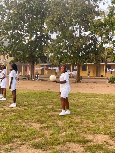 In Emure-Ekiti, Nigeria, on a dusty, outdoor field with sparse grass, a group of young women in white uniforms are engaged in a recreational activity. One woman in the foreground, facing left, is holding a white volleyball. She wears a white t-shirt and white shorts. To her left, three other women in similar white uniforms are standing and facing her. The background reveals a yellow, single-story building with several doors and windows, and laundry hanging on lines in front of it. Trees with green foliage are situated in the background and between the building and the field. Another person is seated on the ground near the building, also in white attire. The lighting suggests it is daytime, likely late afternoon given the soft shadows. The overall scene depicts a moment of leisure or a sports activity at what appears to be an educational or residential institution.